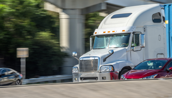 White tractor trailer rig driving under highway underpass with red car on its right.